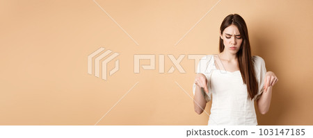 Upset young woman frowning and looking down, pointing at bad thing, standing confused and displeased on beige background Upset young woman frowning and looking down, pointing at bad thing, standing confused and displeased on beige background 103147185