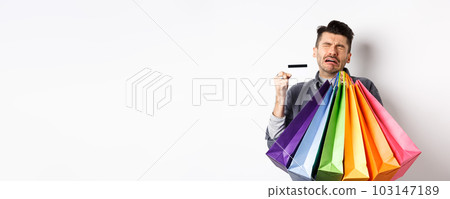 Sad crying man wasted all money on shopping, holding bags and showing empty credit card, standing against white background 103147189