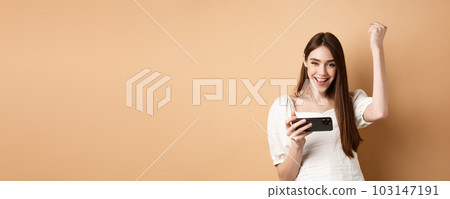 Girl winning on mobile phone. Happy woman raising hand up and scream yes with joy, achieve goal in smartphone app, standing on beige background Girl winning on mobile phone. Happy woman raising hand up and scream yes with joy, achieve goal in smartphone app, standing on beige background 103147191