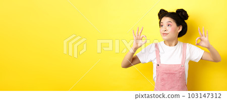 Romantic and beautiful asian girl looking at logo left, showing okay signs in approval, standing on yellow background Romantic and beautiful asian girl looking at logo left, showing okay signs in approval, standing on yellow background 103147312