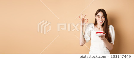 Happy birthday girl show okay gesture and hold bday cake, making wish on her holiday, standing on beige background 103147449