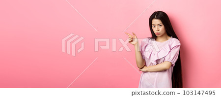 Sad and grumpy asian girl sulking, pointing left and look at camera jealous, want something, complaining and showing desired thing, standing on pink background 103147495