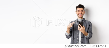 Happy handsome caucasian man with moustache pointing finger at smartphone, smiling and showing good deal online on mobile phone, standing pleased against white background 103147551