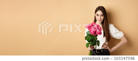 Valentines day. Beautiful girlfriend holding pink roses and looking at camera. Young woman receive flowers from her date, standing on beige background 103147596