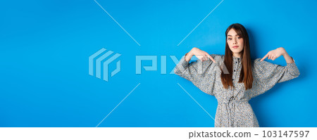 Confident pretty girl in dress pointing fingers at center logo, showing banner and looking self-assured, standing on blue background 103147597