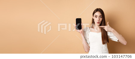Excited woman showing news on screen, pointing at empty phone and looking surprised, standing on beige background 103147706