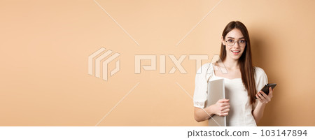 Smiling young woman in glasses reading exciting news on mobile phone, holding laptop and looking happy at camera, standing on beige background Smiling young woman in glasses reading exciting news on mobile phone, holding laptop and looking happy at camera, standing on beige background 103147894