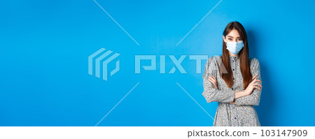 Covid-19, pandemic lifestyle concept. Serious young woman with long natural hair, wearing medical mask, cross arms on chest, protect herself from virus, blue background 103147909