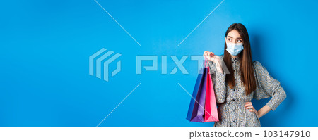 Covid-19, pandemic and lifestyle concept. Girl wearing medical mask on shopping in malls, holding paper bags and look aside at empty space, standing on blue background 103147910