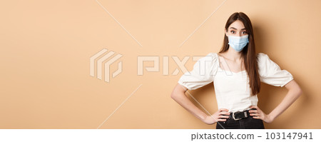 Covid-19 and lifestyle concept. Woman in medical mask looking alarmed at camera, standing on beige background 103147941