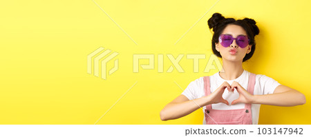 Summer and fashion concept. Cute glam girl with sunglasses and hairbuns, showing heart sign and pucker lips for kiss, I love you gesture, yellow background 103147942