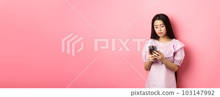 Teenage asian girl chatting on mobile phone, look at smartphone screen with serious face, standing against pink background Teenage asian girl chatting on mobile phone, look at smartphone screen with serious face, standing against pink background 103147992