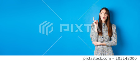 Thoughtful attractive girl in dress pitching an idea, raising finger and looking up, have a plan, standing on blue background 103148000