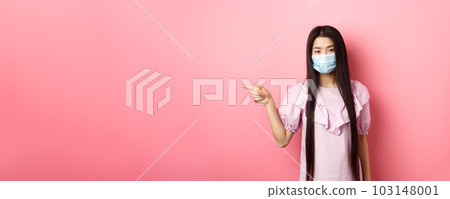 Covid-19, pandemic lifestyle concept. Cute asian teen girl in medical mask pointing finger left at logo, showing advertisement, standing in dress on pink background 103148001