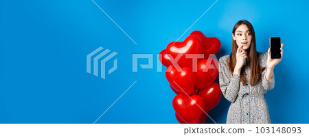 Valentines concept. Beautiful girl looking and showing mobile screen, checking out online shopping offer on lovers day, standing on blue background Valentines concept. Beautiful girl looking and showing mobile screen, checking out online shopping offer on lovers day, standing on blue background 103148093