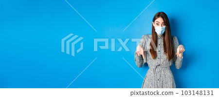 Covid-19, social distancing and healthcare concept. Excited and surprised female model in medical mask, pointing fingers down with disbelief and amazement, blue background Covid-19, social distancing and healthcare concept. Excited and surprised female model in medical mask, pointing fingers down with disbelief and amazement, blue background 103148131