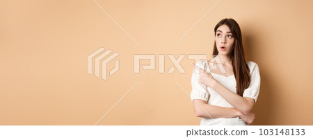 Young woman in dress looking and pointing at upper left corner wondered, saying wow, checking out advertisement with intrigued face, beige background Young woman in dress looking and pointing at upper left corner wondered, saying wow, checking out advertisement with intrigued face, beige background 103148133