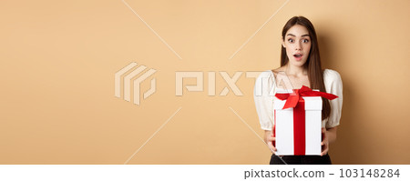 Amazed beautiful girl receiving gift from secret admirer on Valentines day, looking surprised at camera, holding box and standing on beige background Amazed beautiful girl receiving gift from secret admirer on Valentines day, looking surprised at camera, holding box and standing on beige background 103148284
