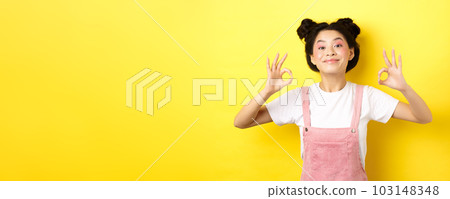 Happy teen koren girl with beauty makeup, showing okay signs and smiling satisfied, recommending product, praise good choice, standing on yellow background 103148348