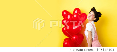 Valentines day and relationship concept. Fashionable asian woman posing near red hearts balloons, surprise for lover on special date, yellow background 103148503
