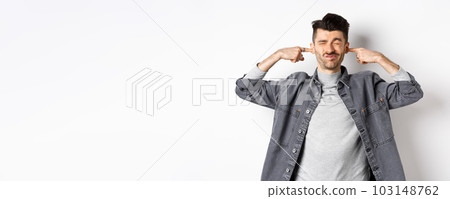 Annoyed young man plug ears with fingers and grimacing from loud noise, disturbed by noisy neighbours, standing at loud place on white background 103148762