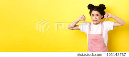 Disgusted asian girl showing thumbs down and cringe from bad product, standing in summer clothes against pink background Disgusted asian girl showing thumbs down and cringe from bad product, standing in summer clothes against pink background 103148787