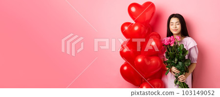 Valentines day concept. Romantic teen asian girl dreaming of love or date, close eyes and smile, holding flowers from lover, receive bouquet of roses and red heart balloons, pink background 103149052