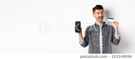 Cheerful handsome guy showing good deal, empty smartphone screen and plastic credit card, winking at you and smiling, recommending offer, white background Cheerful handsome guy showing good deal, empty smartphone screen and plastic credit card, winking at you and smiling, recommending offer, white background 103149096
