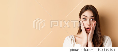 Face of surprised girl saying wow, touching cheeks and staring excited at camera, standing on beige background Face of surprised girl saying wow, touching cheeks and staring excited at camera, standing on beige background 103149104