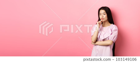 Hmm, interesting. Pensive and cunning asian girl having idea, plan to do something, looking at camera and touching chin with thoughtful face, standing on pink background 103149106