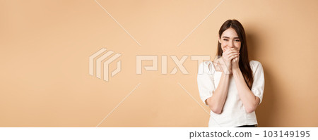Happy girl laughing and covering mouth with hands, hiding her smile and chuckle over something funny, standing on beige background 103149195