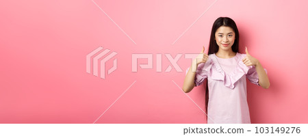 Well done. Smiling asian girl in dress showing thumbs up, like and approve product, praise great deal, standing against pink background 103149276