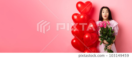 Surprised asian girl in dress standing near valentines day heart balloons and say wow at camera, holding flowers bouquet from lover, romantic date with roses, pink background Surprised asian girl in dress standing near valentines day heart balloons and say wow at camera, holding flowers bouquet from lover, romantic date with roses, pink background 103149459
