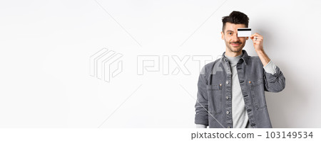 Handsome modern guy with moustache, standing in casual clothes and holding plastic credit card on eye, smiling pleased at camera, white background 103149534