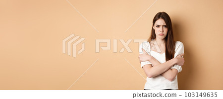 Offended woman hugging herself and frowning upset, feeling timid and defensive, standing on beige background 103149635