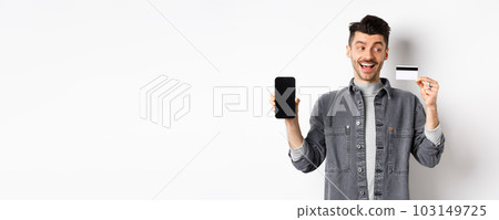 Excited handsome man showing plastic credit card and empty smartphone screen, advertising application, standing on white background 103149725