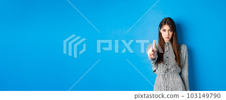 Serious woman looking confident and shaking finger, say no, stop you, disagree and disapprove, standing in dress on blue background Serious woman looking confident and shaking finger, say no, stop you, disagree and disapprove, standing in dress on blue background 103149790