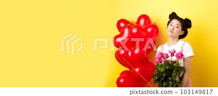 Happy Valentines concept. Sad and gloomy asian woman holding bouquet of roses and feeling upset and lonely on romantic lovers day, standing near red heart balloons, yellow background Happy Valentines concept. Sad and gloomy asian woman holding bouquet of roses and feeling upset and lonely on romantic lovers day, standing near red heart balloons, yellow background 103149817