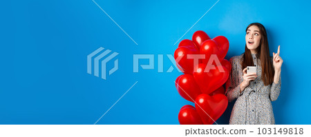 Valentines day. Image of romantic girl pitching an idea after using mobile phone, raising finger up and looking at empty space, standing near red heart balloons, blue background 103149818