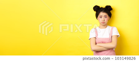 Sad asian teen girl sulking, cross arms on chest, pouting and frowning, complaining on unfair thing, standing against yellow background Sad asian teen girl sulking, cross arms on chest, pouting and frowning, complaining on unfair thing, standing against yellow background 103149826
