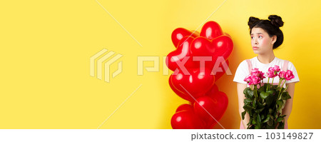 Happy Valentines day. Sad and lonely girl looking left upset, holding bouquet of roses, standing alone near red hearts balloons and frowning, yellow background Happy Valentines day. Sad and lonely girl looking left upset, holding bouquet of roses, standing alone near red hearts balloons and frowning, yellow background 103149827