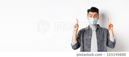 Pandemic lifestyle, healthcare and medicine concept. Excited man advertising in medical mask, pointing fingers up and look amazed, standing on white background 103149880