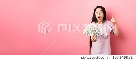 Excited teen girl holding big amount of money, showing dollar bills and thumbs up, standing amazed on pink background Excited teen girl holding big amount of money, showing dollar bills and thumbs up, standing amazed on pink background 103149951