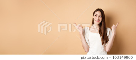 Dreamy caucasian girl making choice, pointing fingers sideways and looking at upper left corner pensive, picking product, standing on beige background 103149960