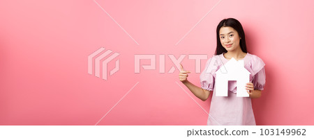 Real estate and insurance concept. Beautiful asian woman pointing at paper house cutout, showing agency logo, standing in dress on pink background 103149962
