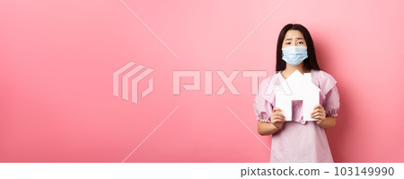 Real estate and pandemic concept. Begging asian girl showing paper house cutout, wearing medical mask, asking for apartment, standing against pink background 103149990