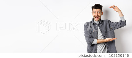 Cheerful guy smiling and showing big size, showing large thing and looking excited, standing against white background Cheerful guy smiling and showing big size, showing large thing and looking excited, standing against white background 103150146