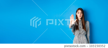 Serious and confident woman with long hair and dress, stretch out hand and say no, make stop gesture, prohibit bad action, standing on blue background Serious and confident woman with long hair and dress, stretch out hand and say no, make stop gesture, prohibit bad action, standing on blue background 103150352
