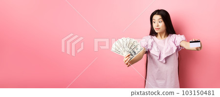Excited teen asian girl stretch out hands with dollar bills and plastic credit card, look at money with amazement, standing on pink background 103150481
