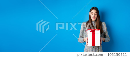 Holidays. Surprised young woman looking at gift with amazed face, receive Valentines day present, standing in dress on blue background Holidays. Surprised young woman looking at gift with amazed face, receive Valentines day present, standing in dress on blue background 103150511